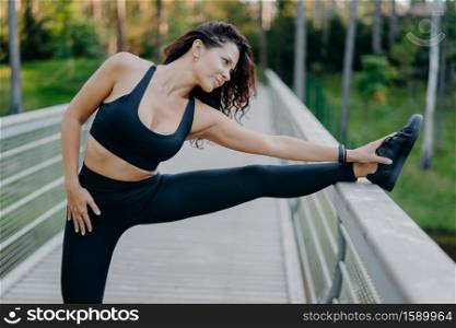 Sporty slim brunette woman dressed in cropped top and leggings stretches legs on bridge does warm up before morning run pose outdoor wants to have slim body and good health. Workout, lifestyle