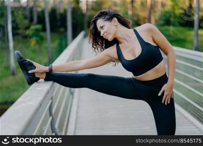 Sporty slim brunette woman dressed in cropped top and leggings stretches legs on bridge does warm up before morning run pose outdoor wants to have slim body and good health. Workout, lifestyle