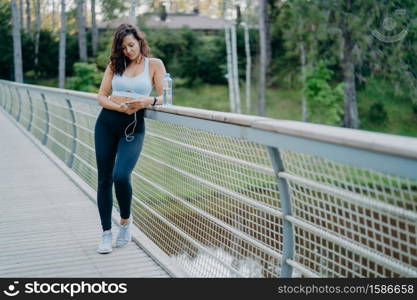 Sporty slim European woman in active wear checks notification on mobile phone, listens music from playlist uses earphones, poses on bridge against nature background. Sport and lifestyle concept