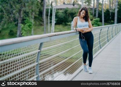 Sporty slim European woman in active wear checks notification on mobile phone, listens music from playlist uses earphones, poses on bridge against nature background. Sport and lifestyle concept