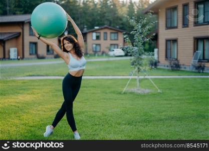 Sporty slim woman wears cropped top and leggings, has aerobics exercises with fitness ball, poses outdoor on green lawn, private houses in background. Healthy fit female model raises fitball over head