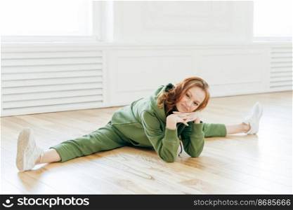 Sporty strong young woman has flexible body does stretching exercise, sits on full split, holds hands together under chin, dressed in tracksuit, poses on floor in empty room for training gymnastics
