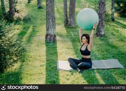 Sporty woman exercises on fit≠ss ball in lotus pose, wearing cropped top and≤ggings, in the forest during a sunny day. Yoga practice, fresh air.