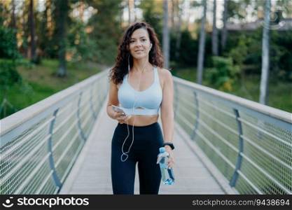 Sporty woman in active wear jogs near forest, listens to music, holds water bottle, poses on bridge. Outdoor fitness concept.