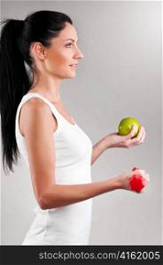 sporty woman is holding red barbell and green apple on grey background