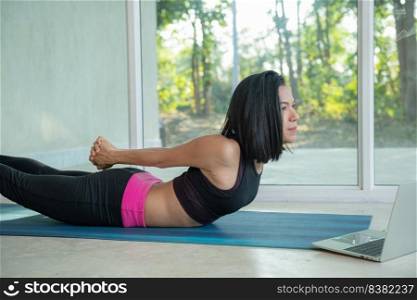 Sporty woman practicing yoga, back and buttock training pose,stretching watching fitness video tutorial online on laptop, doing workout at home sitting on mat in living room practicing