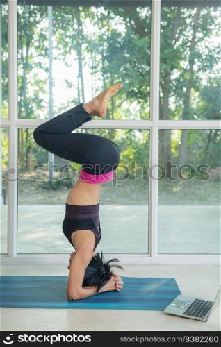 sporty woman practicing yoga, doing headstand exercise, salamba sirsasana pose, working out, wearing sportswear black, watching fitness video tutorial online on laptop, doing workout at home sitting.