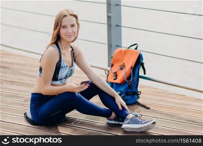 Sporty woman wears casual top, leggings and sportshoes, looks positively at camera, holds cell phone in hands, listens radio online with earphones, has break after outdoor training. Pastime concept