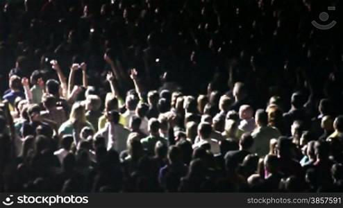 Spotlights illuminate sections of a crowd of cheering fans at a rock concert.