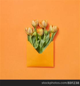 Spring fresh flowers tulips in craft envelope on a yellow background with copy space. Love letter for congratulation. Flat lay. Post card with bouquet of tulips in craft envelope on an orange background.