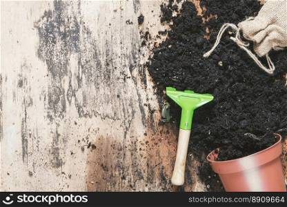 Spring gardening theme image with an overturned flower pot full of soil, a jute sack with seeds and a green rake on a vintage wooden table.