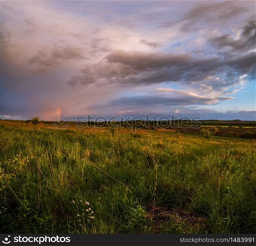 Spring meadow after rain, cloudy evening pre sunset sky with rainbow, rural hills and fields in far. Natural seasonal, weather, climate, countryside beauty concept scene.