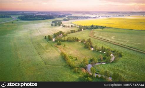 Spring morning aerial rural panorama. Sunrise over green and yellow blooming colza fields. Clouds of fog and small river with trees on riverbank. Belarus, Europe