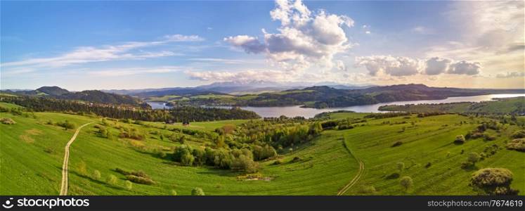 Spring mountain panorama. Lake Czorsztyn on Dunajec river. Village in green forest and meadow hills. Cloudy evening sunset sky. Aerial view of Tatra range woodland landscape. Poland, Malopolska region