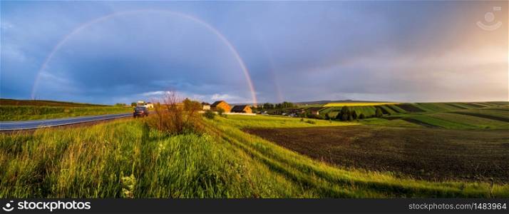 Spring rapeseed and small farmlands fields after rain evening view, cloudy pre sunset sky with colorful rainbow and rural hills. Natural seasonal, climate, farming, countryside beauty concept scene.