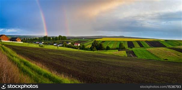 Spring rapeseed and small farmlands fields after rain evening view, cloudy pre sunset sky with colorful rainbow and rural hills. Seasonal, weather, climate, eco, farming, countryside beauty concept.