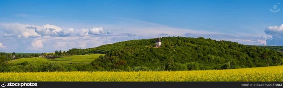 Spring rapeseed yellow blooming fields view, blue sky with clouds in sunlight panorama. Pyatnychany tower  defense structure, 15th century  on far hill slope.