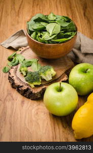 Spring spinach leaves in the bowl, broccoli, lemons and apples on wooden table background