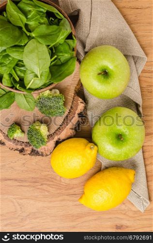 Spring spinach leaves in the bowl, broccoli, lemons and apples on wooden table background