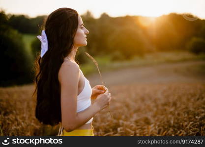 spring, sunset, wheat field, girl, evening, sun, yellow skirt, white t-shirt, white bow, hand with a spike, rustic style, girl&rsquo;s smile, bread field, model, contour light. A girl in a yellow skirt stands in a field against the background of the setting sun