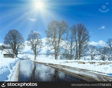 Spring sunshine and road through the alpine village in Austria with reflection of trees in thawing snow.