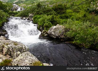 Spring waterfall in mountain valley. Fast river with green shores