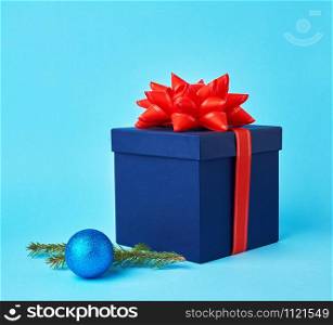 square blue cardboard box with a red bow, sprig of spruce with a shiny blue ball on a blue background, festive Christmas background. Christmas greeting card