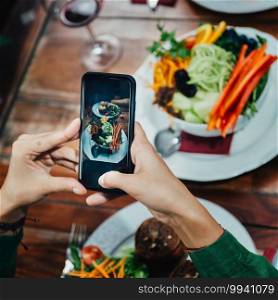 Square image of young woman photographing delicious vegetarian burger and mixed organic salad on white plate in vegetarian restaurant.