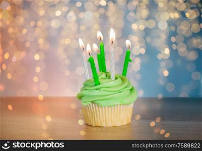 st patricks day, birthday, holidays and celebration concept - green cupcake with six burning candles on table over festive lights. green cupcake with six burning candles on table