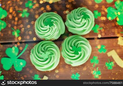 st patricks day, food and holidays concept - green cupcakes and shamrock on wooden table top view over festive lights. green cupcakes and shamrock on wooden table