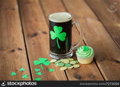 st patricks day, holidays and celebration concept - glass of dark draft beer with shamrock, green cupcake and gold coins on wooden table. shamrock on glass of beer, green cupcake and coins