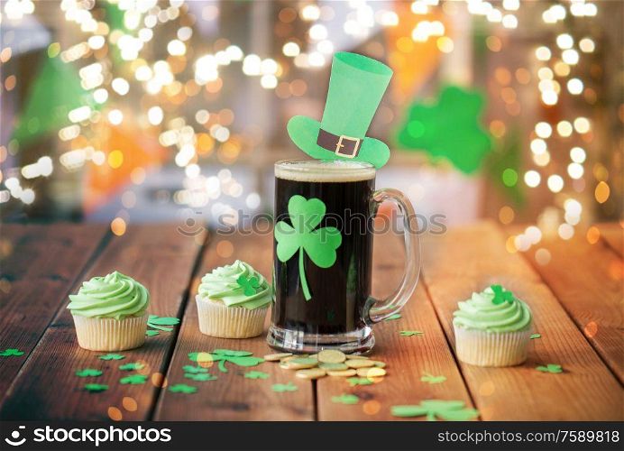 st patricks day, holidays and celebration concept - glass of dark draft beer with shamrock, green cupcakes and gold coins on wooden table over festive lights. shamrock on beer glass, green cupcakes and coins