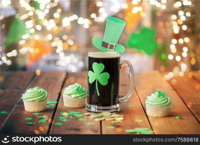 st patricks day, holidays and celebration concept - glass of dark draft beer with shamrock, green cupcakes and gold coins on wooden table over festive lights. shamrock on beer glass, green cupcakes and coins