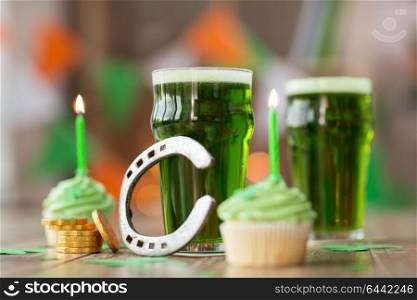 st patricks day, holidays and celebration concept - glass of green beer, cupcakes with candles, horseshoe and gold coins on table. glass of beer, cupcakes, horseshoe and gold coins