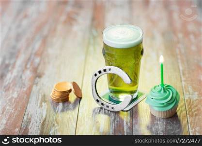 st patricks day, holidays and celebration concept - glass of green beer, cupcake with candle, horseshoe and gold coins on wooden table. glass of beer, cupcake, horseshoe and gold coins