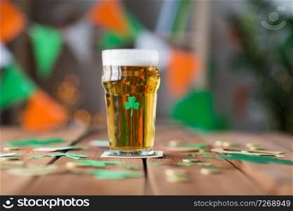 st patricks day, holidays and celebration concept - glass of light beer with shamrock decoration and gold coins on wooden table. glass of beer with shamrock and coins on table