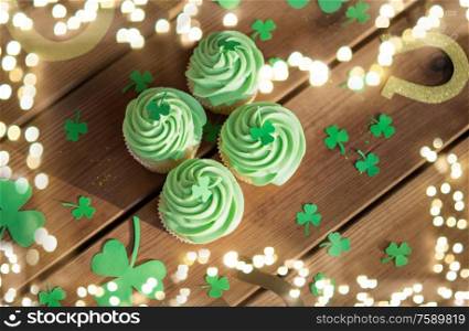st patricks day, holidays and celebration concept - green cupcakes, horseshoes and shamrock on wooden table over festive lights. green cupcakes, horseshoes and shamrock