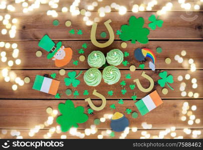 st patricks day, holidays and celebration concept - green cupcakes with candles, horseshoes and shamrock on wooden table over festive lights. green cupcakes and st patricks day decorations