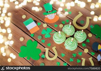 st patricks day, holidays and celebration concept - green cupcakes with candles, horseshoes and shamrock on wooden table over festive lights. green cupcakes and st patricks day decorations