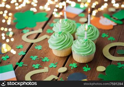 st patricks day, holidays and celebration concept - green cupcakes with candles, horseshoes and shamrock on wooden table over festive lights. green cupcakes and st patricks day decorations