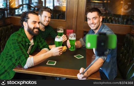 st patricks day, leisure and technology concept - happy male friends drinking green beer and taking picture with smartphone selfie stick at bar or pub. friends taking selfie with green beer at pub