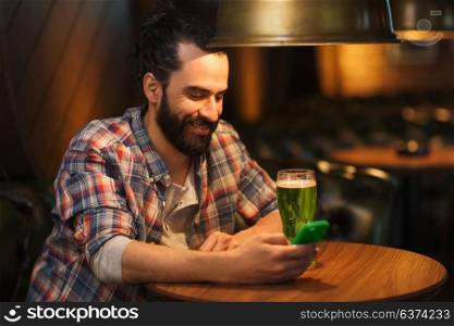 st patricks day, people and technology concept - happy man with smartphone drinking green beer and reading message at bar or pub. man with smartphone and green beer texting at bar