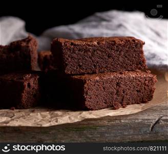 stack of baked square pieces of chocolate brownie cake on brown parchment paper, black background