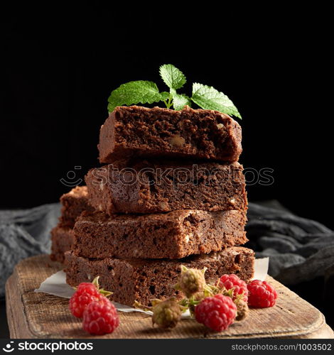 stack of baked square pieces of chocolate brownie cake on brown wooden cutting board, black background