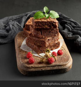 stack of baked square pieces of chocolate brownie cake on brown wooden cutting board, black background
