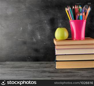 stack of books and multi-colored wooden pencils, background of an empty black chalk board, back to school concept