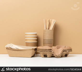 stack of brown disposable paper cups and a tray on a white table, wooden forks and knives, brown background. Utensils for takeaway drinks, zero waste