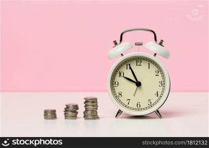 stack of coins round white alarm clock on the table. The concept of time is money, a period of discounts.