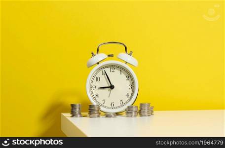 stack of coins round white alarm clock on the white table. The concept of time is money, period of discounts.
