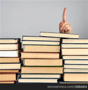 stack of different books on a gray background, a hand sticks out from behind a pile and shows up the index finger, attention sign
